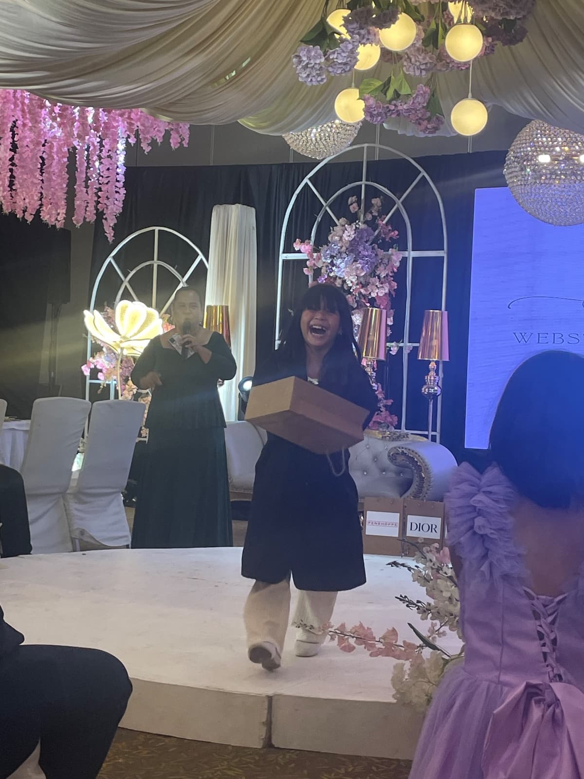 A guest walking back to her seat mid-laugh with the prize box, host mid-announcement behind her.