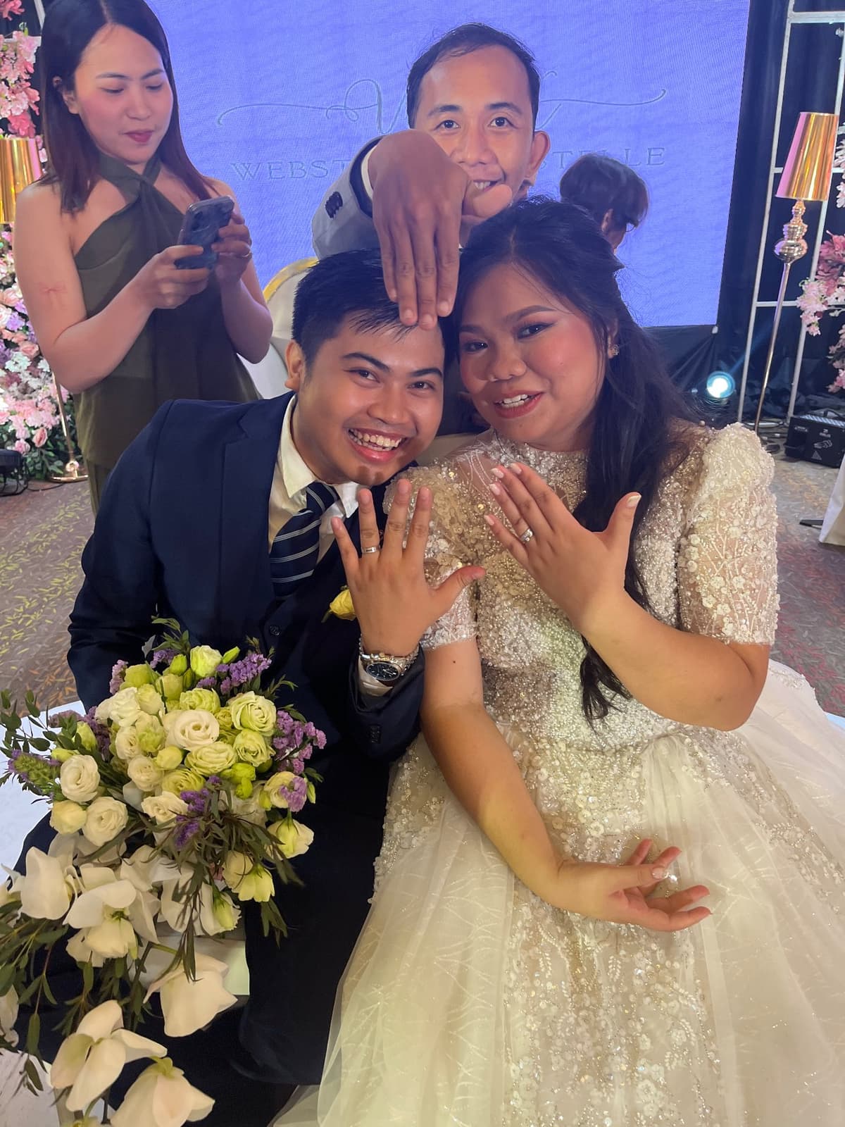 Webster and Krystelle showing their rings mid-celebration while a guest behind them snaps a photo on her phone.
