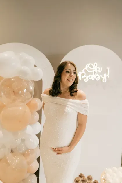 A pregnant woman in a white dress cradles her belly beneath a balloon arch and an “Oh Baby” neon sign.