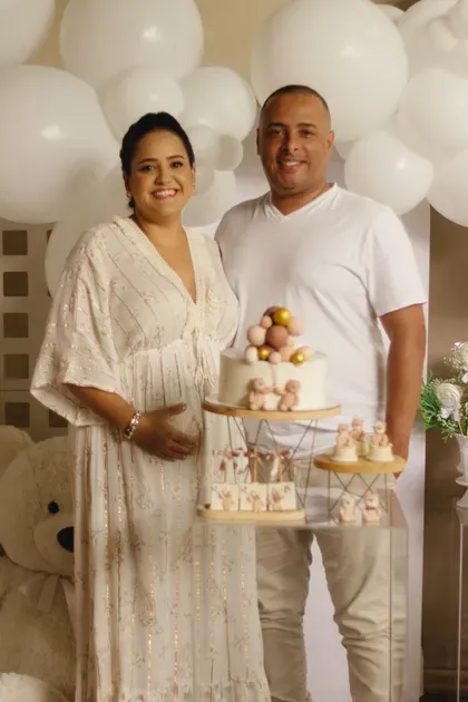 An expectant couple hold a small cake in front of a soft cream balloon backdrop.