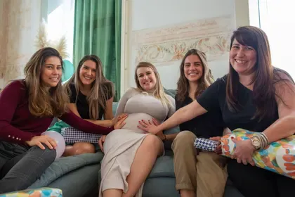 Friends on a sofa rest their hands gently on the mom-to-be’s belly beside a teddy bear.