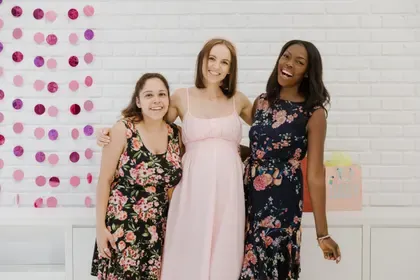 Three friends stand close together laughing, the pregnant guest in blush pink at the centre.