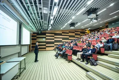 A seated audience watches a speaker beside a large projection screen at a conference.